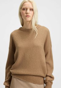 Brauner Strickpullover mit rundem Ausschnitt und überschnittenen Schultern, der eine dezente Textur und einen gerippten Saum aufweist, sowie ein kleines Logo-Detail am unteren Rand.