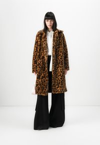 Kvinne med svart hår som har på seg en brun leopardmønstret pelsejakke, hvit bluse, vide svarte bukser og holder en lys rosa håndveske.