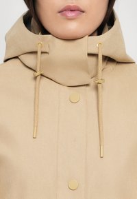 Veste beige à capuche avec quincaillerie dorée, cordons de serrage et boutons-pression. Tissu doux et texturé avec un design simple et fonctionnel.