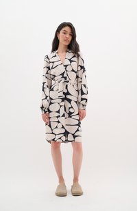 Long-sleeve wrap dress in cream and black abstract pattern, featuring a belt, a v-neckline, and puffed sleeves. Worn with beige slip-on shoes.