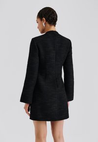 Schwarzes, strukturiertes Kleid mit langen Ärmeln und einem hohen Ausschnitt. Das Kleid hat eine taillierte Silhouette und eine Nahtdetail, die entlang des Rückens verläuft.