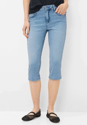 Pantalon capri en denim bleu clair avec poches avant cloutées, porté avec des chaussures plates en mesh noir et un haut noir, coupé juste en dessous du genou.