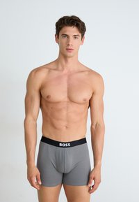 Boxer-briefs gris avec une ceinture élastique noire portant le logo "BOSS". Design ajusté, tissu doux, texture lisse. Pas de détails supplémentaires.