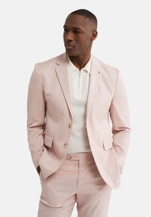 Man draagt lichtroze pak met bijpassende broek en witte gebreide trui met ritssluiting aan de voorkant, handen in de zakken, kijkt lichtjes opzij.