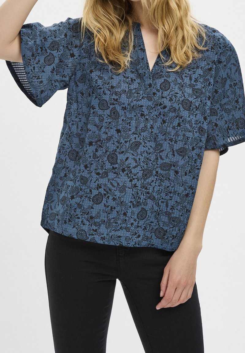 Top stampato floreale blu in tessuto leggero, con maniche corte, scollatura a V e vestibilità ampia. Contrasto di colore tra nero e blu.