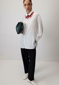 Witte overhemd met knoopsluiting en een rode kralenketting, zwarte broek en een groene leren clutch. Het model draagt witte schoenen en staat tegen een neutrale achtergrond.