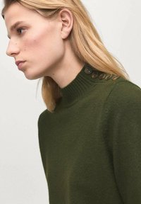 Maglione verde a maglia con collo alto, dotato di bottoni marroni sulla spalla. Texture morbida, design aderente e dettagli minimi.