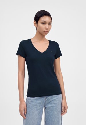 Jeune personne aux cheveux courts et foncés portant un t-shirt sombre à col en V et un jean bleu clair sur un fond clair uni.