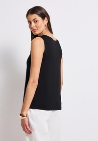 Czarny bezrękawnik z okrągłym dekoltem, wykonany z miękkiego materiału, z prostym dołem, zestawiony z białymi spodniami i złotą bransoletką na nadgarstek.
