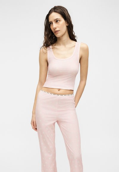 Γυναίκα με μακριά καστανά μαλλιά, φοράει ανοιχτό ροζ αμάνικο crop top και διαφανές πουά παντελόνι που ταιριάζει, στέκεται με το ένα χέρι στην τσέπη.