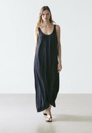 Frau geht vorwärts, trägt ein ärmelloses schwarzes Maxikleid und schwarze Zehensandalen vor einem schlichten hellen Hintergrund.