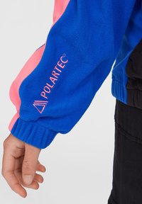 Blå fleecejakkeærme med lyserøde detaljer, der har et broderet "POLARTEC"-logo. Ribbet manchetstruktur.