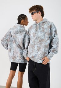 Nadrozmerné mikiny s mramorovým vzorom v šedej a béžovej farbe, so predným vreckom a kapucňou na sťahovanie, v kombinácii s čiernymi šortkami a džínsami.