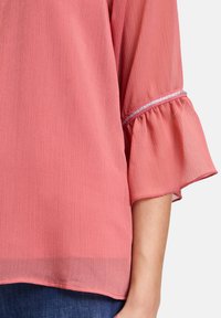 Blusa leggera di colore rosa con tessuto texturizzato. Presenta maniche svasate con un accento argentato a contrasto sull'orlo.