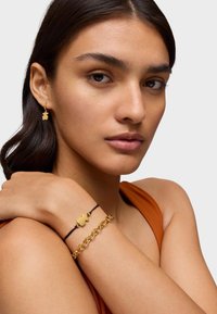 Pendientes de aro de oro y una pulsera con capas que incluye un charm de oro, en un modelo que lleva una blusa naranja. Se aprecian texturas de metal suave y cuero.