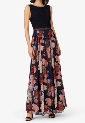 Mouwloze jurk met zwarte kanten top en lange donkerblauwe rok met grote roze en rode bloemmotieven, gedragen met zwarte open-teen hakken.