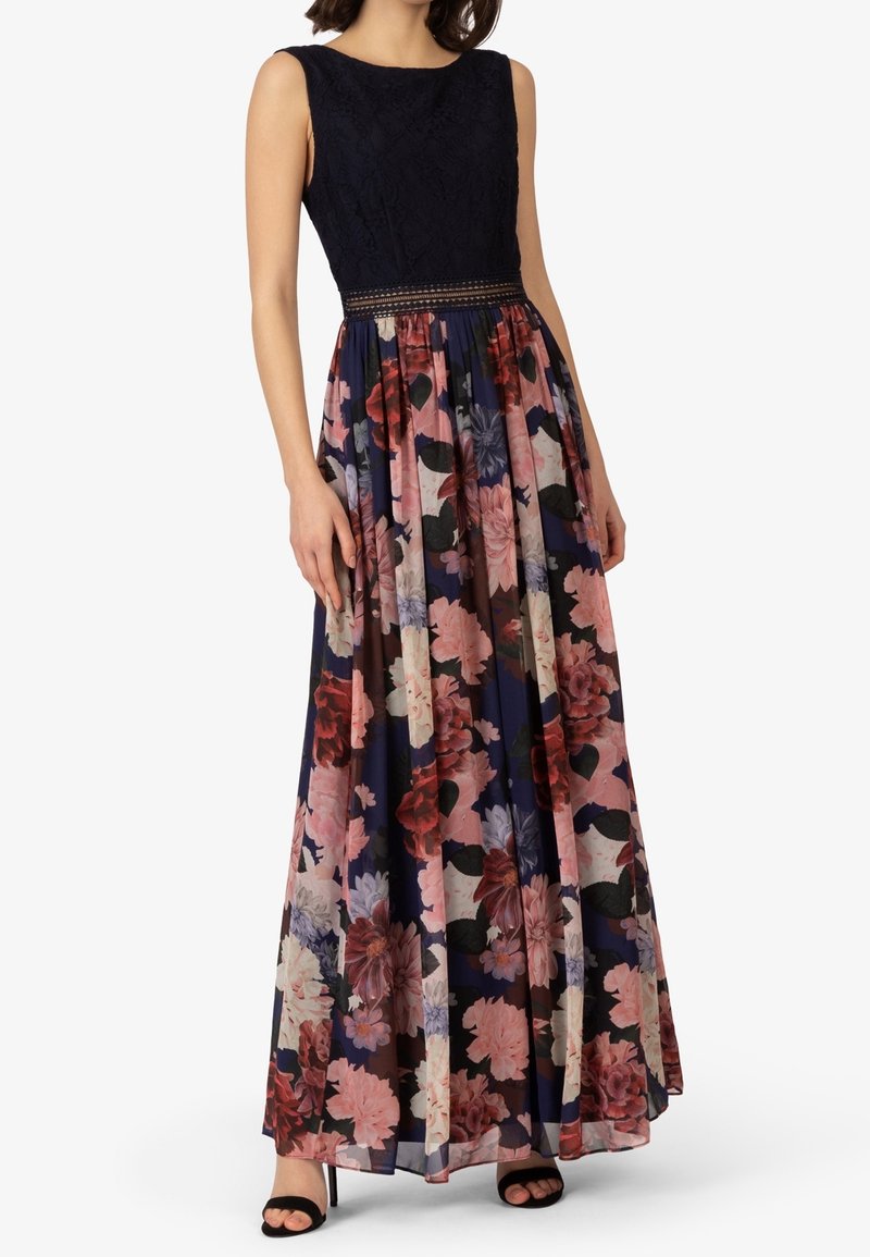 Robe sans manches avec un haut en dentelle noire et une longue jupe bleu marine ornée de grands motifs floraux roses et rouges, portée avec des talons ouverts noirs.