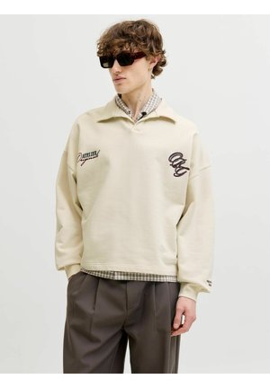 Noor mees tumedate päikeseprillidega, beež pullover trükitud tekstiga, all kandes ruudulist särke ning tumedat halli volditud pükste vastu ühevärvilist tausta.