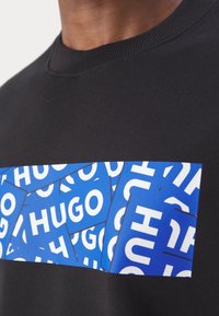 Gros plan d’un t-shirt noir à col rond avec des patches rectangulaires bleus et blancs superposés épelant « HUGO » en lettres épaisses.