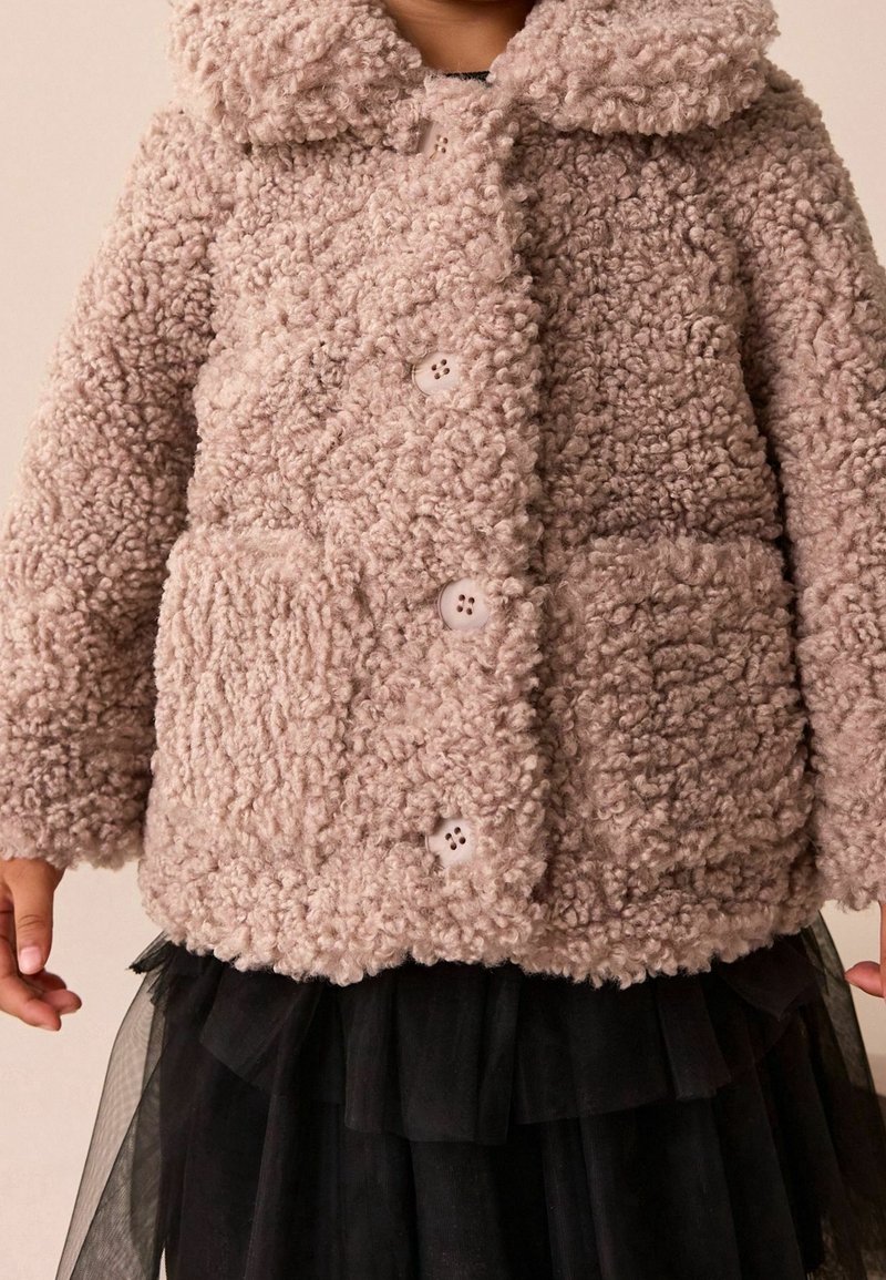Enfant portant une veste en laine beige texturée avec des boutons et des poches avant sur une jupe noire à couches.