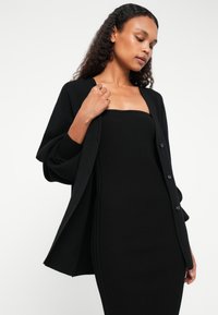 Μαύρο πλεκτό cardigan με χαλαρή εφαρμογή και υπερμεγέθη μανίκια, που διαθέτει μπροστινά κουμπιά. Φορεμένο πάνω από ένα μαύρο ριμπ φόρεμα με αντιστοιχία.