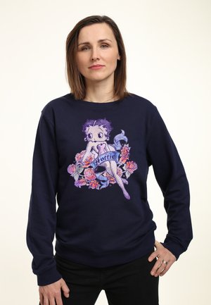 Sweatshirt bleu marine présentant une impression graphique d'un personnage assis entouré de roses colorées, avec le mot "Hottie" sur un ruban en dessous.