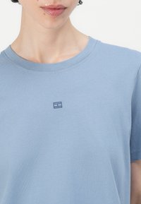 Šviesiai mėlynos medvilninės marškinėliai su apvalia iškirpte ir trumpomis rankovėmis; širdies srityje yra mažas tamsiai mėlynos spalvos išsiuvinėtas logotipas.