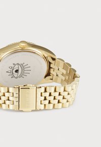 Bracelet et fermoir en métal doré avec une gravure "TIMEX", montrant l'arrière d'un boîtier de montre rond présentant un design graphique d'œil.