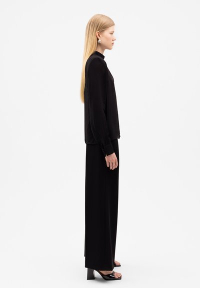 Schwarzes langärmliges Oberteil und passende weit geschnittene Hose, mit einer glatten Textur und hohem Kragen. Die Accessoires umfassen große Creolen.