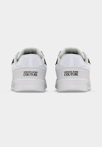 Ζευγάρι λευκά sneakers Versace Jeans Couture, εμφανιζόμενα από πίσω, με μαύρο λογότυπο στα ταμπελάκια της φτέρνας, πάνω σε απλό γκρι φόντο.