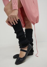 Femme ajustant des chauffe-jambes noirs à côtes sur des ballerines noires, portant une robe rose avec un ourlet à cordon.