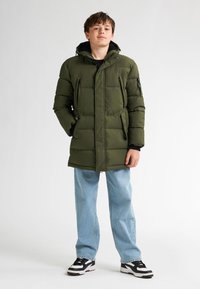 Olivgrüne Steppjacke mit einem Reißverschluss und Knopfverschluss, mehreren Taschen und Kapuze. Kombiniert mit hellblauen Jeans und schwarzen Sneakers.