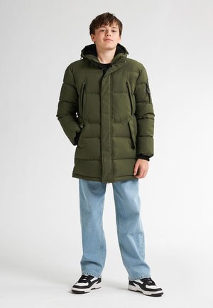 Olijfgroene pufferjas met een rits- en knoopsluiting, meerdere zakken en een capuchon. Gecombineerd met lichtblauwe spijkerbroeken en zwarte sneakers.