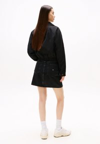 Sort denimnederdel med lige snit og baglommer, kombineret med en afslappet sort jakke. Modellen har hvide sneakers og sokker på.