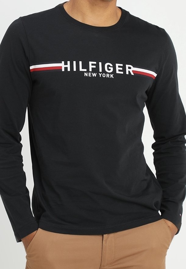 Mężczyzna w czarnej koszuli z długim rękawem z logo "HILFIGER NEW YORK" oraz czerwonymi i białymi pasami na klatce piersiowej, widoczne beżowe spodnie.