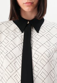 White blouse with black geometric pattern, black collar, and a vertical tie. Features a small gold button at the collar. Smooth texture.