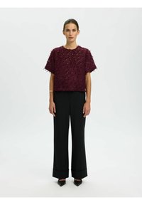 Bordeaux textuur top met losse pasvorm, korte mouwen en franjes, gedragen met zwarte wijde broek en scherpe zwarte schoenen.