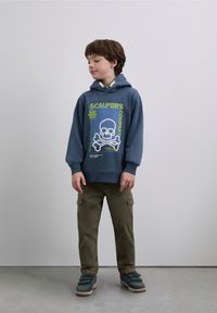 Felpa con cappuccio blu scuro con grafica frontale, raffigurante un teschio con ossa incrociate in bianco e verde. Abbinata a pantaloni cargo oliva e sneakers verdi.