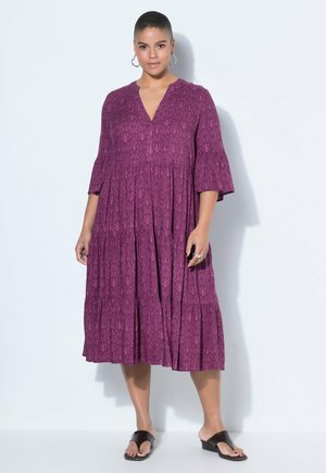 Kvinde med kort hår iført en lilla midi-kjole med mønster, klokkeærmer og sorte hælede sandaler, stående mod en hvid baggrund.