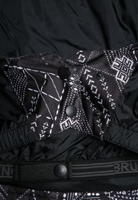 Tissu noir avec des motifs géométriques blancs, muni de boutons-pression noirs et d'une ceinture élastique portant partiellement le texte "BRU".