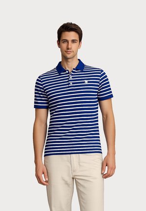 Man draagt een blauw-wit gestreept poloshirt met korte mouwen en een kraag, gecombineerd met lichtbeige broek, staand tegen een effen achtergrond.