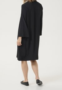 Robe noire en tissu lisse, avec de longues manches larges, une coupe ample et une longueur jusqu'aux genoux. Portée avec des chaussures plates noires.