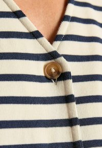 Pruhovaný pletený kardigán v krémovej a námorníckej modrej farbe, s výstrihom do V a okrúhlym béžovým gombíkom pri golieri. Textúra je hladká a mäkká.