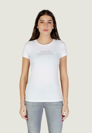 Giovane donna che indossa una t-shirt aderente bianca Armani Exchange e jeans grigio chiaro, in piedi contro uno sfondo semplice.