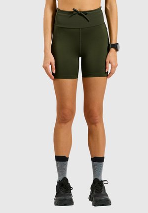 Woman wearing olive green high-waisted athletic shorts, gray crew socks, black running shoes, and a black wristwatch standing against a white background.
