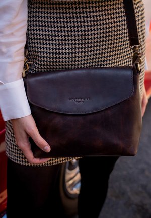 Brown leather shoulder bag with a smooth texture, a flap closure, and a logo. Worn over a houndstooth-patterned outfit.