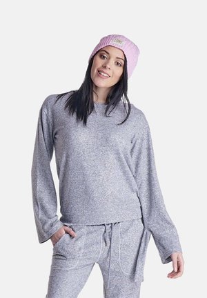 Jeune femme portant un haut gris ample à manches longues, un pantalon assorti avec un cordon et un bonnet rose en tricot, debout avec une main dans la poche.
