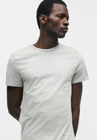 Άνδρας με σκουρόχρωμο δέρμα που φοράει απλό ανοιχτό γκρι κοντομάνικο μπλουζάκι, κοιτάζοντας προς τα πλάγια μπροστά σε μονόχρωμο λευκό φόντο.