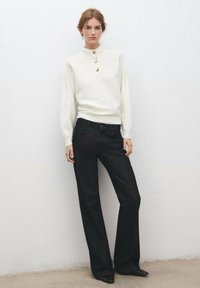 Femme debout contre un mur blanc, portant un pull blanc à manches longues avec des boutons dorés et un pantalon large noir avec des chaussures noires pointues.