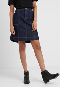 Falda de mezclilla oscura con diseño en línea A, bolsillos en la parte delantera, acentos de costura blanca y dobladillo a media longitud. Llevada con botas de tobillo negras.